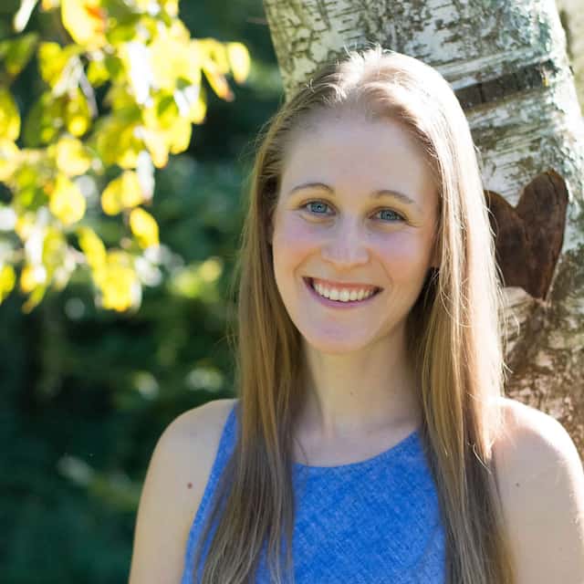 Woman with long blonde hair smiling in front of a tree trunk with a heart carved into it, surrounded by green foliage